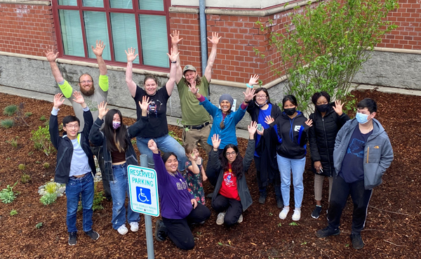 A group of 13 people smile at the camera and hold up their hands waving. They are standing or kneeling in the rain garden.