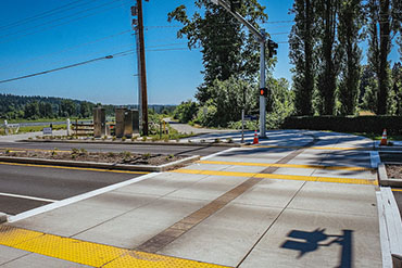 A wide crosswalk, complete with tactile paving and protective median, crosses a two-lane road to connect to a paved, tree lined trail on the other side of the street.